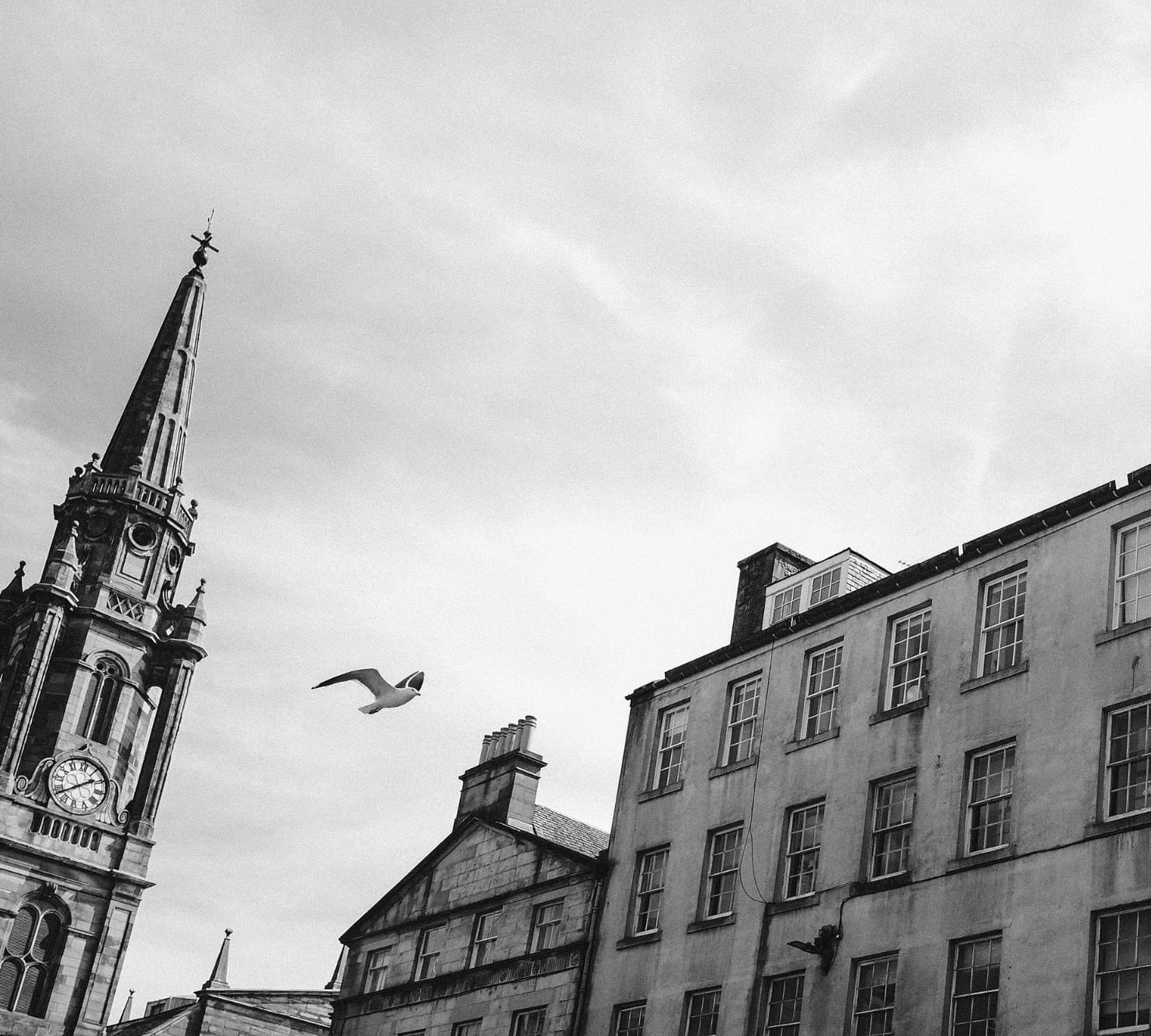 Black and white photograph of a church spire and stone buildings against a cloudy sky with a bird in flight