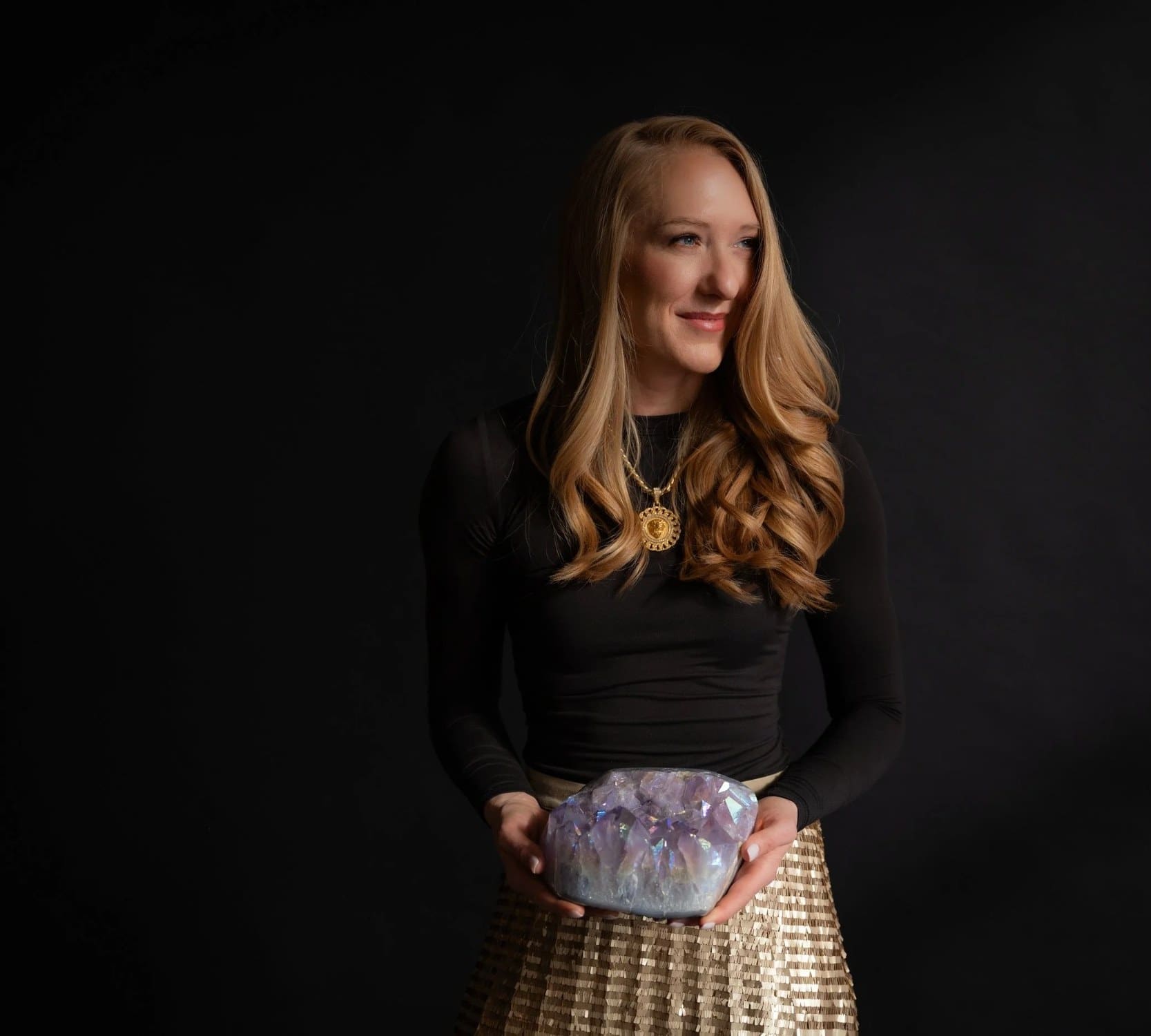 A woman with long wavy hair in a black top and gold sequin skirt holding a purple amethyst geode against a dark backdrop
