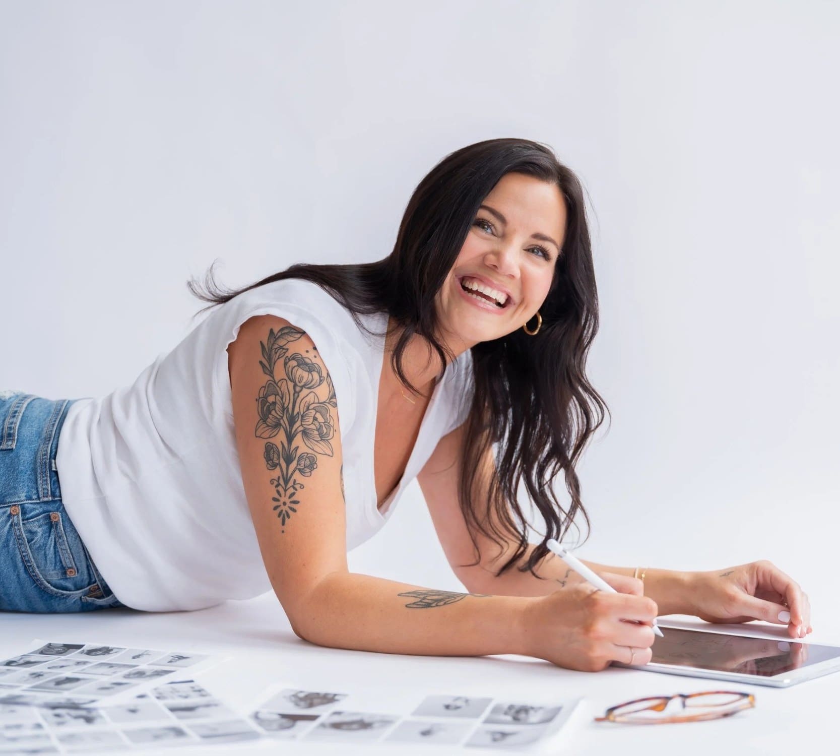 Smiling dark-haired woman with an arm tattoo lying on a white studio floor sketching on a tablet beside contact sheets