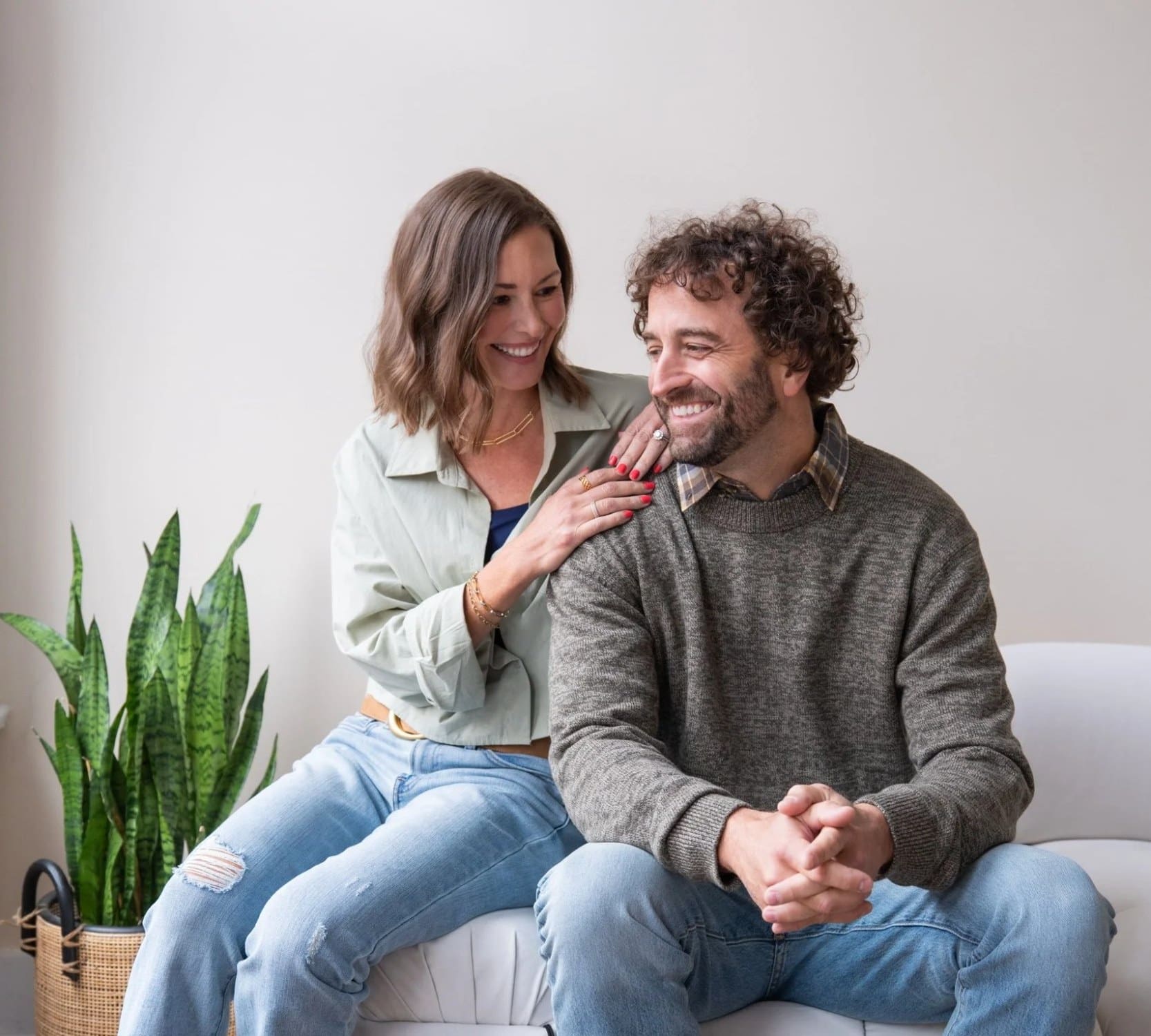 A woman in a sage green jacket and a man in a gray sweater laughing together on a white sofa during a branding session