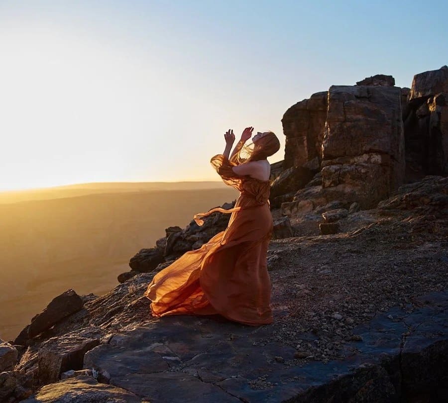 Woman in a flowing orange dress with her arms raised at sunset on a rocky cliff in the Namibian desert
